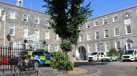 The courtyard of the Guernsey Police HQ. The three-storey buildings are grey stone and there is a glasshouse style entrance. There are police cars and van parked in front and there is a tree and some iron railings in the foreground.