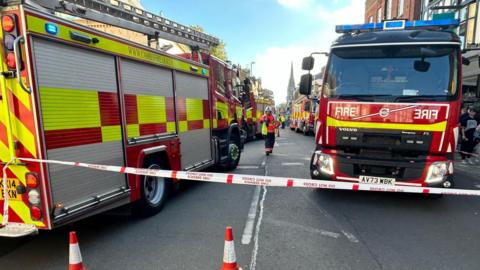 Two fire engines and a firefighter can be seen behind a cordon tape.