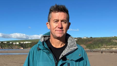 A man in a turquoise changing robe with a black t-shirt underneath stands on a beach in November sun.