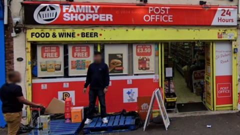 Two men unloading pallets of goods outside a local shop called Family Shopper. It also serves as a post office.