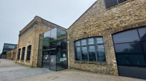 Sand Martin House is a one storey bricked building which has large black panned windows. In the middle there is an entrance made from glass with a door.