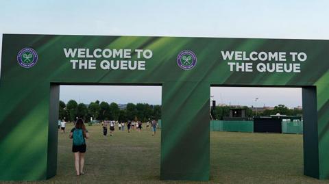 A green entry way which reads: "Welcome to the queue" in large white capital letters. Beyond there is a field, temporary fencing and people walking away from the camera.