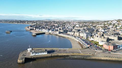 An view of the seaside town of Broughty Ferry. The sea is on the left side of the picture, and the town is on the right, with hills visible in the distance. In the foreground, a pier stretches out into the water. 