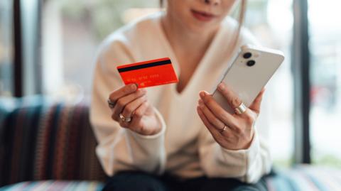 Midsection of young Asian woman holding a credit card while using smartphone, sitting on sofa. Online shopping and electronic payment.