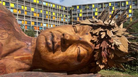 The face of a large wooden sculpture lies on the grass in-front of a huge colourful block of flats. 