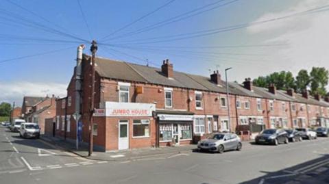 A row of terraced buildings with cars parked on the roadside. There is a junction to the side leading on to another road. One of the buildings has a white sign outside with orange writing which reads 'Jumbo House'.