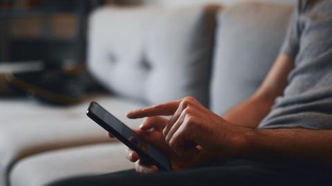 In this stock image, an unidentifiable man sits on a grey sofa, swiping on his smartphone.