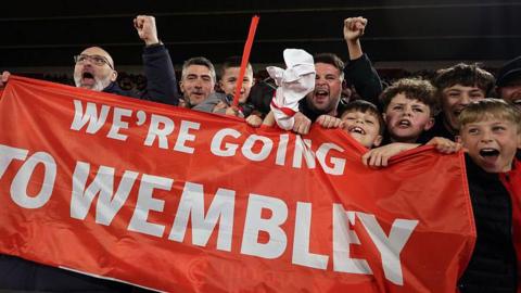 A group of male Southampton football fans holding a red and white banner that says 'we are going to wembley' they are a mix of ages and are all cheering.