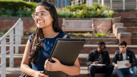 A female student with long straight brown hair and glasses smiles off camera as she stands in front of steps holding a leather folder - behind her, two male students sit on the steps in conversation
