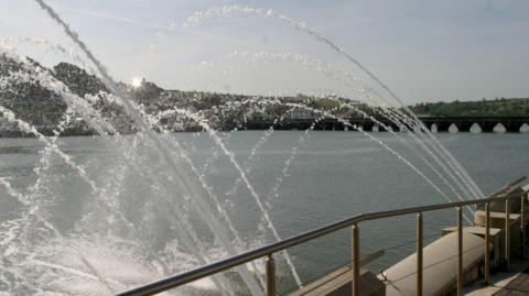 A water fountain spraying water into a river. In the background is a bridge. There is a hill with houses. The sky is a light blue with the sun coming out from behind a house. The river is a dark blue. The water coming out of the fountain is clear. 