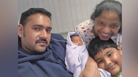 Two adults, one child and a baby posing for a selfie. The man has black hair and facial hair and is holding a tiny newborn baby which is wrapped in a lilac blanket and has a tube coming out of his nose. Leaning his head near the baby is a young boy with black hair and a yellow spongebob squarepants top on. Above him is a woman with black hair and white top which has red roses on it