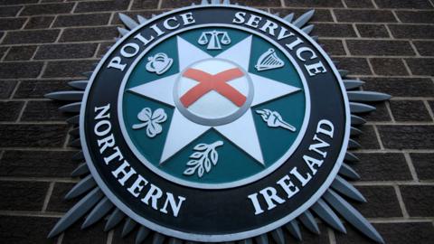 A PSNI crest on a brown brick wall. The crest is a circular white, green and black badge, outlined by a silver star. 