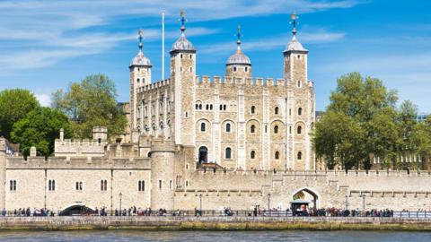 The Tower of London viewed from across the River Thames, showing its stone walls, corner towers with domed roofs, and surrounding grounds under a blue sky, with people walking along the riverside.