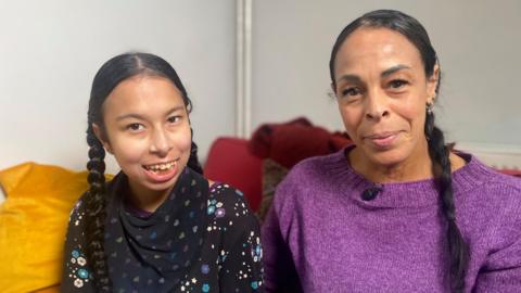 Neave and Natasha are sitting indoors on a sofa. Both have long, dark hair styled in braids/pigtails. Natasha, who is taller, is wearing a purple knitted top with a small microphone clipped near the neckline. Naeve is dressed in a black top patterned with colourful stars and flowers. They are both looking at the camera and smiling.