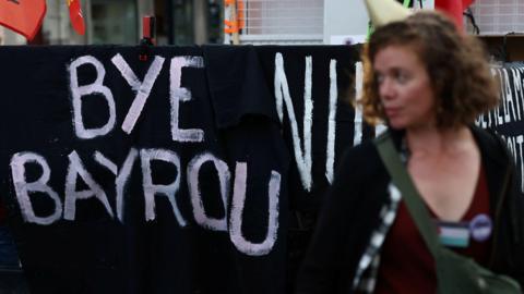 The slogan "bye bayrou" is painted on a black sheet outside France's national assembly as a woman looks on.