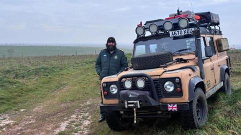Man standing outdoors alongside mustard-coloured Range Rover with camping equipment attached. It is parked grass in a rural area. He has on a dark green ambulance jacket and a black beanie hat, and has a large, dark beard.