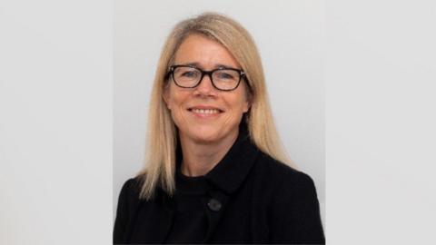 A woman wearing a black coat and black glasses. She is smiling and has shoulder length blonde hair. Behind her is a grey background.