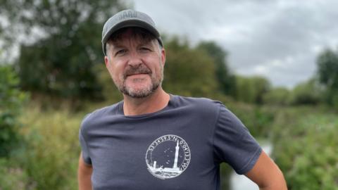 A man with a greying beard stands on the banks of the River Freshney in Grimsby. He is wearing a t-shirt and a baseball cap.