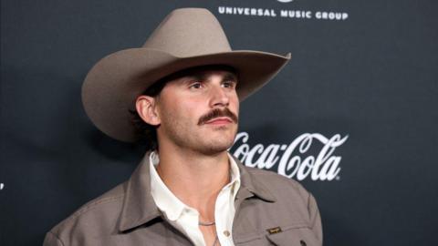 A man with a thick brown moustache and a brown cowboy hat is looking to the right of the camera. He is wearing a white shirt and silver chains and a brown jacket. 