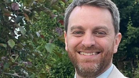 A headshot of a man stood in a garden area, he has short cropped greying hair and a greying beard, and is smiling with his teeth showing. He is wearing a light coloured suit jacket and shirt. 