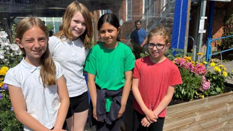 The four girls are pictured on their school yard in front of flower beds. They are all smiling looking at the camera. Seren, on the left, is wearing a white t-shirt and has long blonde hair, which is draping on her left shoulder. Next to her is Eleanor, who again is wearing a white t shirt, black shorts and has light brown shoulder-length hair. Fatima is standing next to Eleanor. She is wearing a green  School t-shirt and has a blue jumper tied around her waist. Fatima has black hair in a ponytail, with a middle parting. Ada, on the right, is wearing a red School t-shirt. She wears glasses and has light brown hair which is in a ponytail.