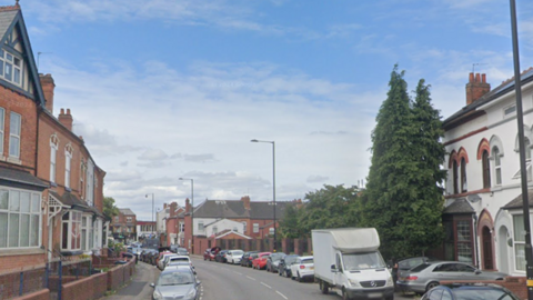 A road with terraced houses and parked cars on either side. The cars range in colour from white to red to black and there is also a white van in among them. The homes are made of brick with bay windows and small front gardens with brick walls at the front.