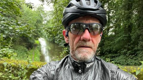 John Ireson with his cycling gear on. He is wearing a helmet and glasses. There is a waterway behind him and lots of trees. 