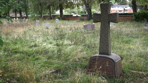 A gravestone in a cemetery, with other gravestones to the left. There is a lot of overgrown grass around the graves and trees and a brick wall in the distance. A building is also behind the graves. 