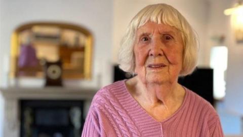 91-year-old Betty Brown with short grey hair, wearing a pink knit top in her living room, with a blurred mantelpiece, fireplace and mirror seen behind her