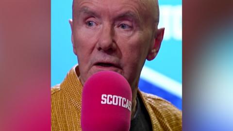 A man wearing a yellow shirt speaks into a pink microphone