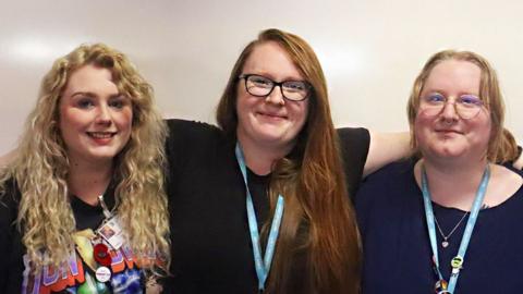 Three women stand against a beige wall with their arms around each others' shoulders. On the left, Charlotte has her blonde curly hair down and she is wearing a black band T-shirt. In the middle, Laurie has her red hair done and on one side and is wearing a black T-shirt and glasses. Stephanie is wearing a navy blue top and heart necklace, and pale glasses. They are all looking at the camera and smiling.