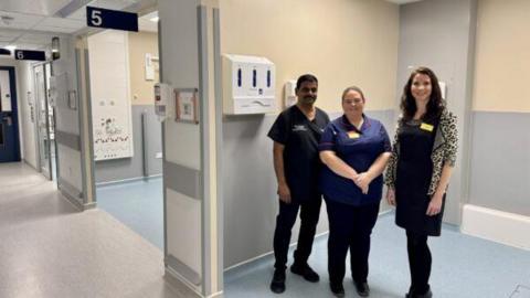 Three people stand in a hospital, in a large open cubicle. A female clinician stands in the middle, wearing trousers and a navy hospital shirt, a man on her left in black scrubs. A woman on the right with long brown hair wears a leopard-print cardigan.