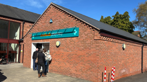 A brick building with the words Rowlands Pharmacy on a mainly green sign above a door. Three people are outside the door.