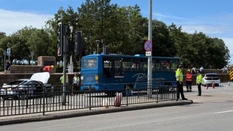 A car with its bonnet up is stationary behind a blue bus and another car is stationary in front of the bus. There are officers in high vis on the street and some cones on the road.