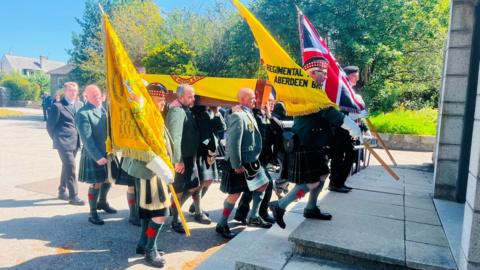 Coffin of veteran Jim Glennie arriving at funeral service, carried by men in kilts, under a blue sky.