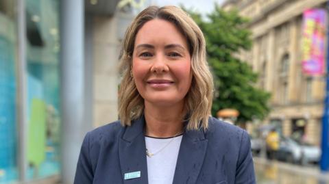 Joanne Briggs has shoulder-length light brown hair and is wearing a white top under a navy jacket. She is wearing a gold "Dad" necklace.