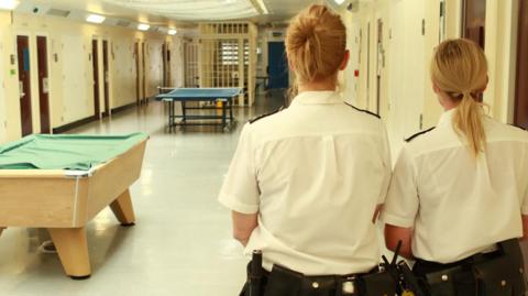 Two female prison officers with their backs to the camera standing an a recreation area of HMP Norwich. A table tennis table and a pool table can be seen. 