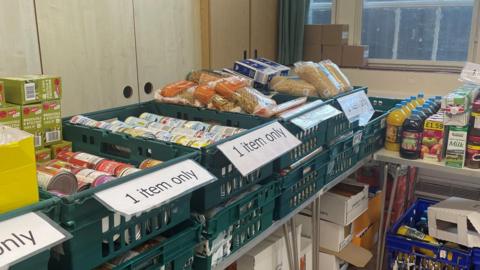 Trestle tables with crates of non-perishable foods. Cans and packets of pasta, fruit juices and milk.