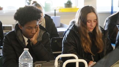 In a classroom, two students sit in their coats while working. One male student with black hair is leaning on his hand while in a puffer jacket. He is sat next to a female student who has long mousey brown hair who is writing with a pen on paper and is wearing a parker style coat.