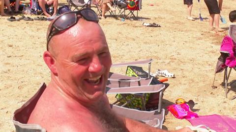 A man is sat in a camping chair laughing. He is wearing sunglasses on top of his head, in the background is sand from the beach and legs of other beach-goers that are not fully in frame.