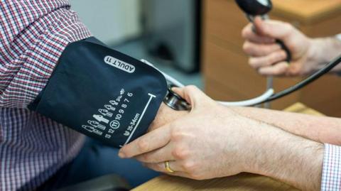 Stock close-up photo of an arm wearing a black blood pressure monitor. The arm has a red and white chequered shirt. The blood pressure is being taken by another arm which is holding down on the blood pressure monitor. 