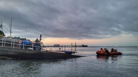 An orange RNLI lifeboat heading out to sea from a concrete slipway. Three RNLI members can be seen on the boat with helmets and life vests. A man sits on a blue tractor halfway up a ramp leading into the sea, he looks back at the lifeboat. The sun is setting in the background behind grey clouds, and Penarth Pier can be seen in the background. 