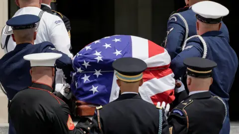 Men in military uniform are seen from behind carrying a casket with a US flag draped over it