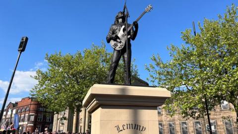 The cast bronze statue showing a man with a guitar in front of a microphone. It stands on a stone plinth with Lemmy written on it. There are buildings and trees surrounding it.
