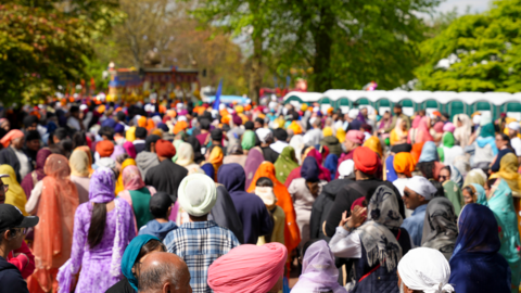 A crowd of tens of thousands of people in a park dressed in brightly coloured clothing facing away from the camera. In the background there appears to be a coloured stage