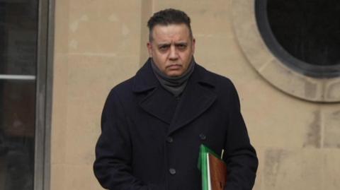 Shahid Malik pictured leaving court. He is wearing a navy overcoat and carrying some files.