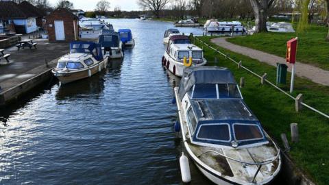 Boats are seen in the foreground with a photograph looking along the River Waveney in Beccles, with clear skies in the background