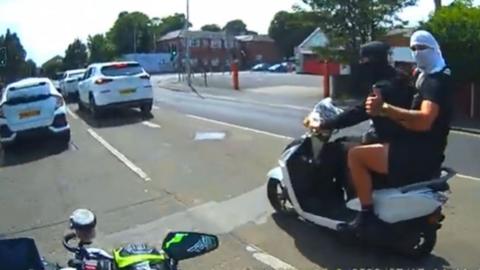 Two males on a white moped pull up alongside a man on a motorbike at traffic lights. The rider is wearing a black jumper and black face covering, the passenger has a black t-shirt and is wearing a white face covering.