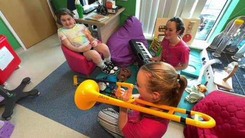 Hephzibah sits on the floor with a keyboard on her knee. Her colleague sits  next to her playing a yellow plastic trombone. A young girl sits in a small pink chair smiling.