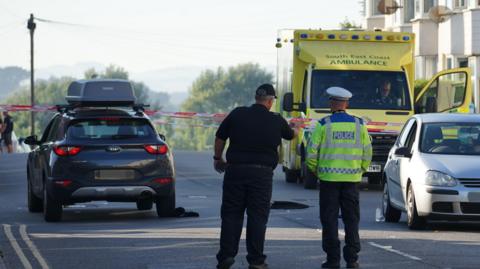 Two police officers stood in a road, facing away from the camera. There is a dark grey vehicle to their left and an ambulance and smaller light grey car to their right